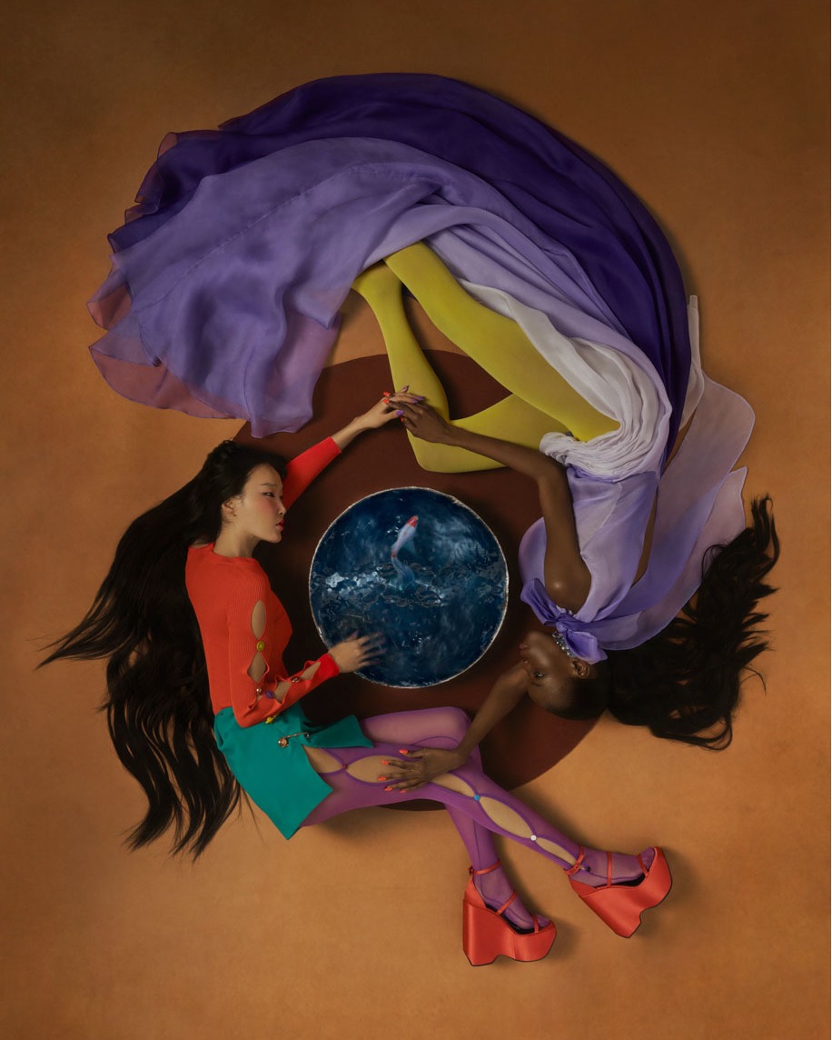 Two models lie in a circle around a fishbowl, wearing orange platform heels, yellow tights, and purple dresses, against a brown backdrop.