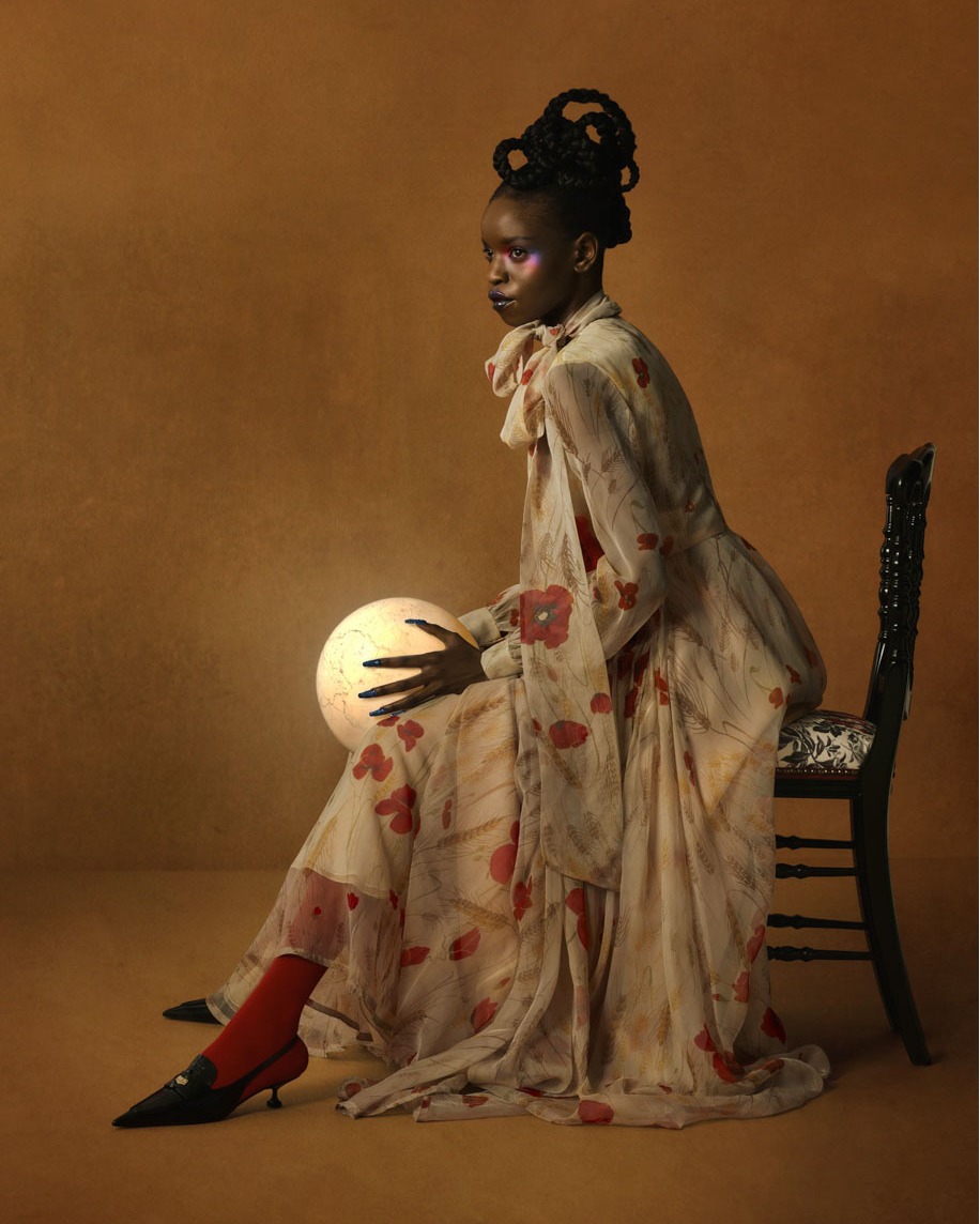 Elegant woman with unique braided hair and an off-white floral dress poses while seated, holding a glowing sphere in a warm-toned studio.