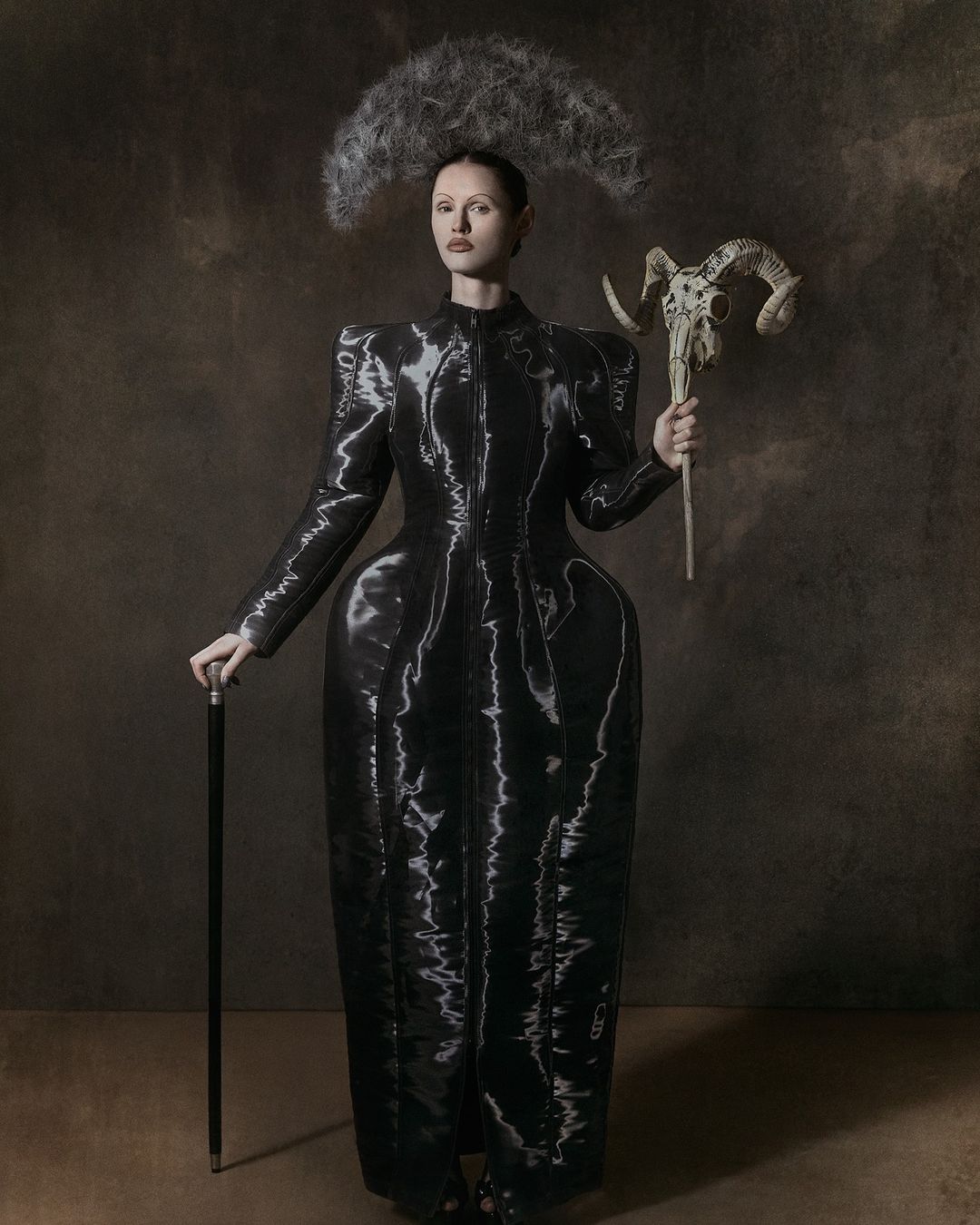 A woman stands in a dark studio wearing a black, futuristic dress with a horned skull staff and abstract headpiece for a fashion editorial.