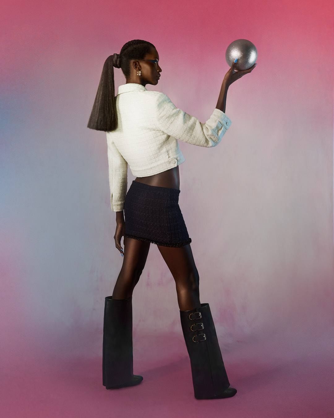 A slender Black woman in a cropped white jacket, black mini skirt, and tall black boots poses holding a metallic silver ball.