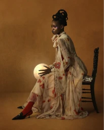 Black woman in a patterned dress holding a glowing orb while seated on a chair against a warm brown textured studio backdrop.