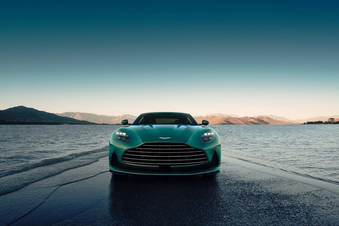Front view of a teal Aston Martin luxury car parked on a wet concrete surface near water, mountains and a light blue sky.