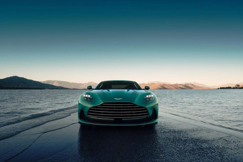 Front view of a teal Aston Martin luxury car parked on a wet concrete surface near water, mountains and a light blue sky.