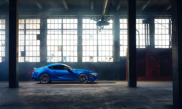 Blue sports car parked inside a large warehouse with bright sunlight streaming through the windows, creating dramatic lighting and shadows.