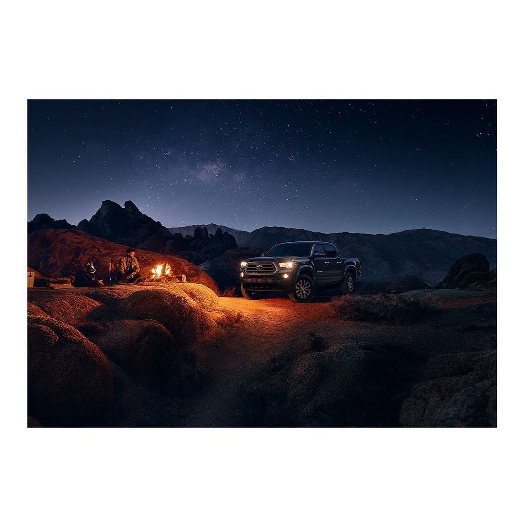 Black pickup truck parked in a desert landscape at night with a campfire, mountains, and starry sky creating a scenic camping view.