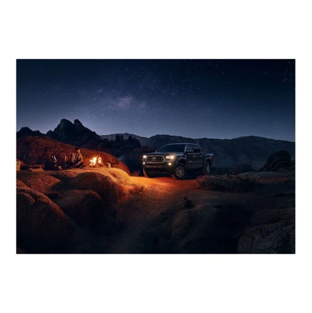 Black pickup truck parked in a desert landscape at night with a campfire, mountains, and starry sky creating a scenic camping view.
