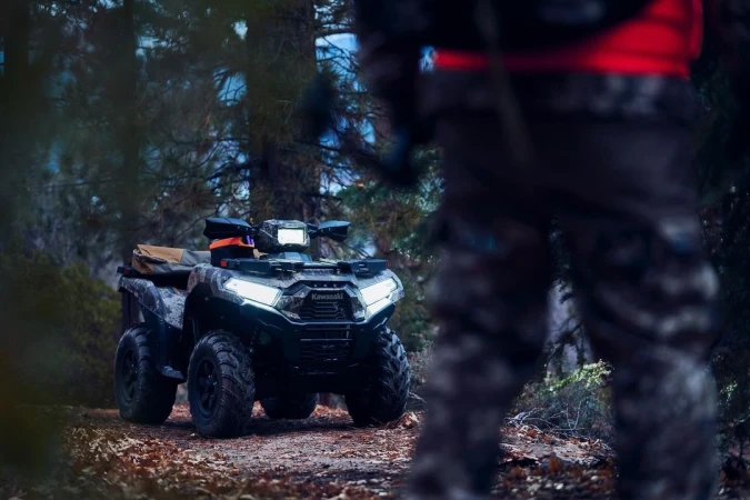 Camouflage Kawasaki ATV parked on a wooded trail with a blurred figure standing nearby, surrounded by trees and fallen leaves.
