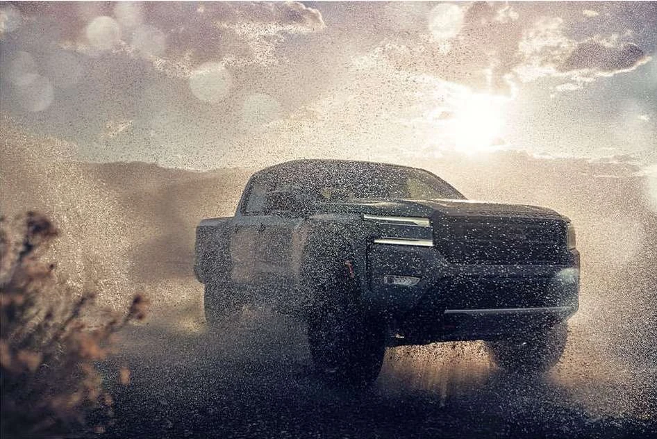A dark grey pickup truck kicks up water on a dirt road against a backdrop of a cloudy sky and distant mountains during daytime.
