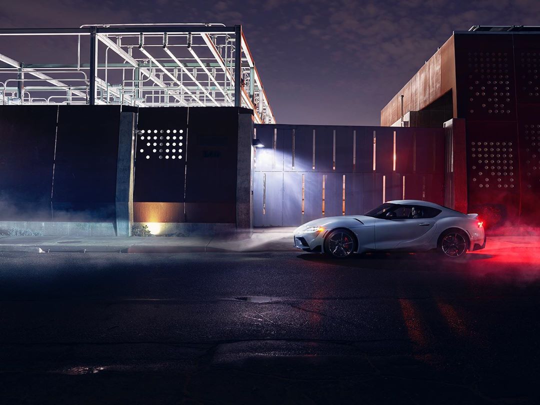 Silver sports car parked on dark asphalt in an urban setting at night, with industrial architecture and glowing lights in the background.