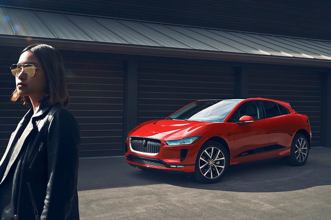 A red Jaguar I-PACE parked in front of a building with an adult female in a black leather jacket and sunglasses standing nearby.