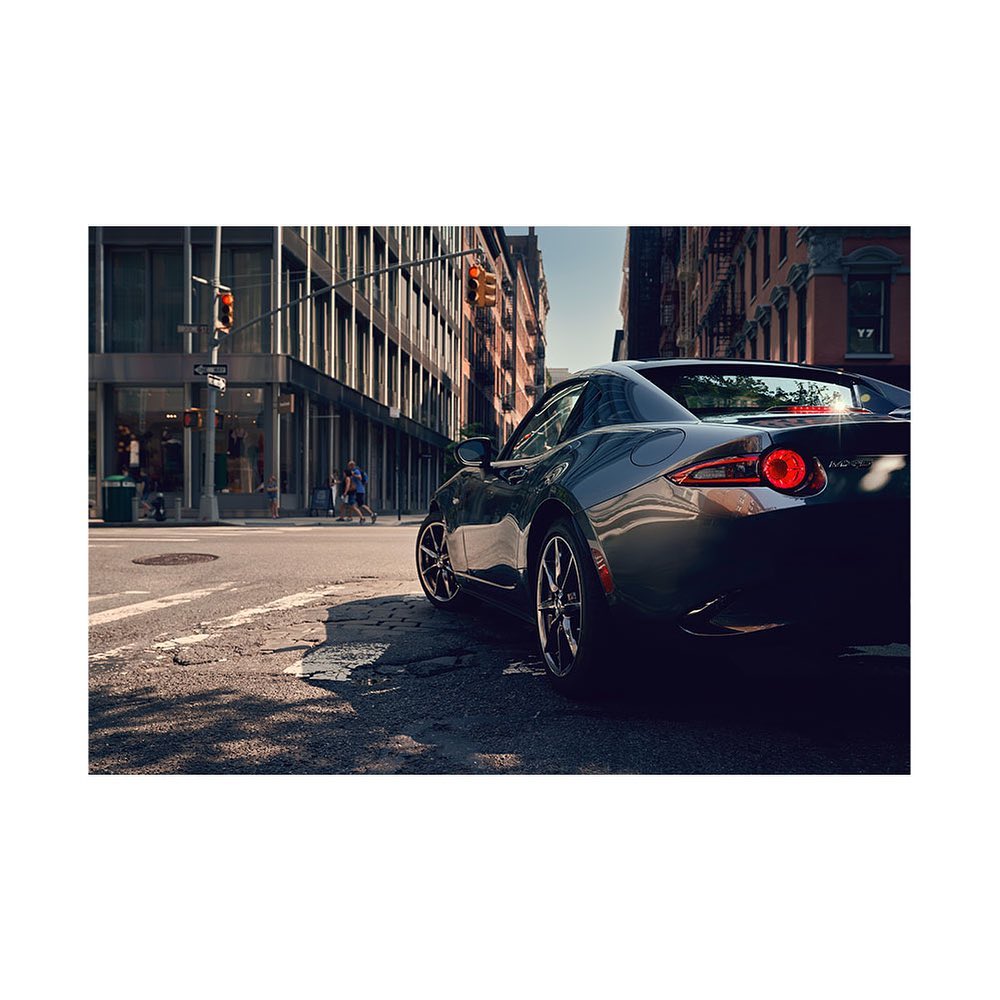 Gray Mazda Miata parked on a city street with buildings and a traffic light in the background, showcasing urban automotive photography.