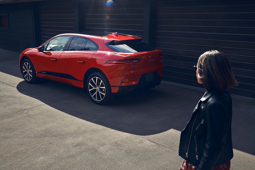 Red Jaguar I-Pace electric vehicle parked near a dark gray building with an adult woman standing beside it, looking at the car.