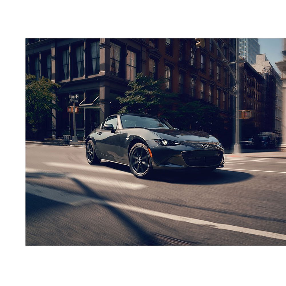 Gray Mazda Miata speeding on a city street with tall buildings, trees, and motion blur creating a sense of velocity and energy.