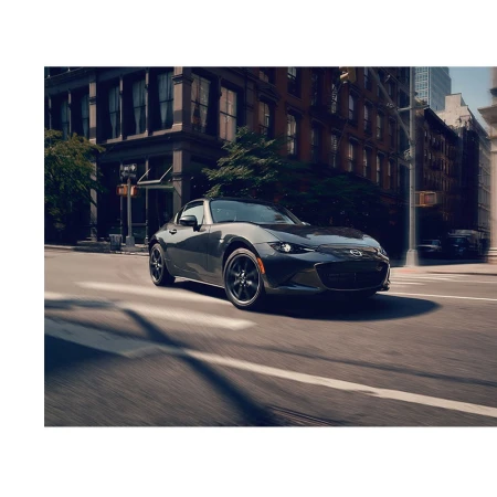 Gray Mazda Miata speeding on a city street with tall buildings, trees, and motion blur creating a sense of velocity and energy.
