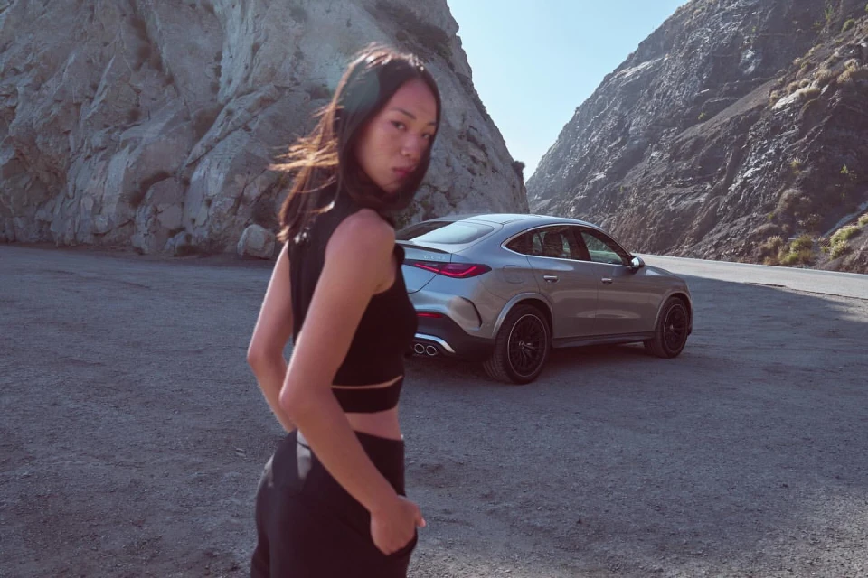 Adult woman posing near a silver SUV on a mountain road. The car is parked on the roadside next to a steep rocky mountain.