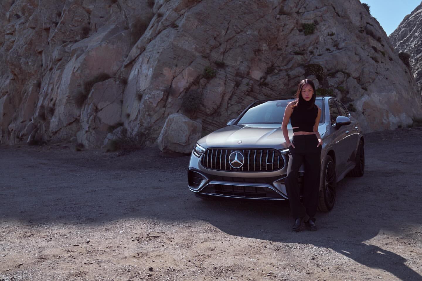 A silver Mercedes-Benz SUV is parked in a rocky desert landscape, with an adult woman posing next to the vehicle.