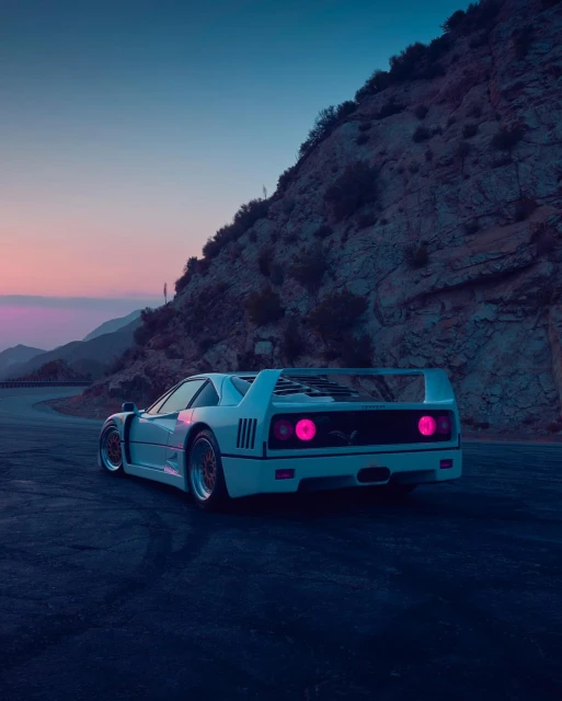 Rear view of a white Ferrari F40 parked on an asphalt road with a mountain and soft twilight sky in the background.
