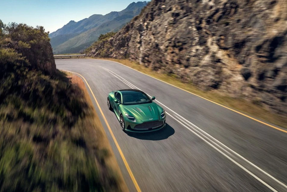 A sleek green Aston Martin DB12 coupe races along a winding mountain road, framed by verdant hills and clear blue skies. 