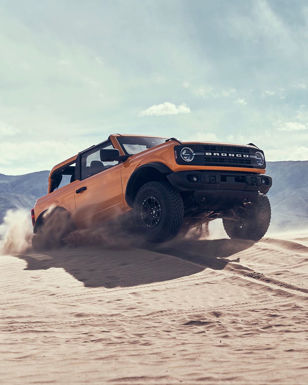 A vibrant orange Ford Bronco SUV drives across a sandy dune under a partly cloudy sky, kicking up sand in a dynamic outdoor shot.