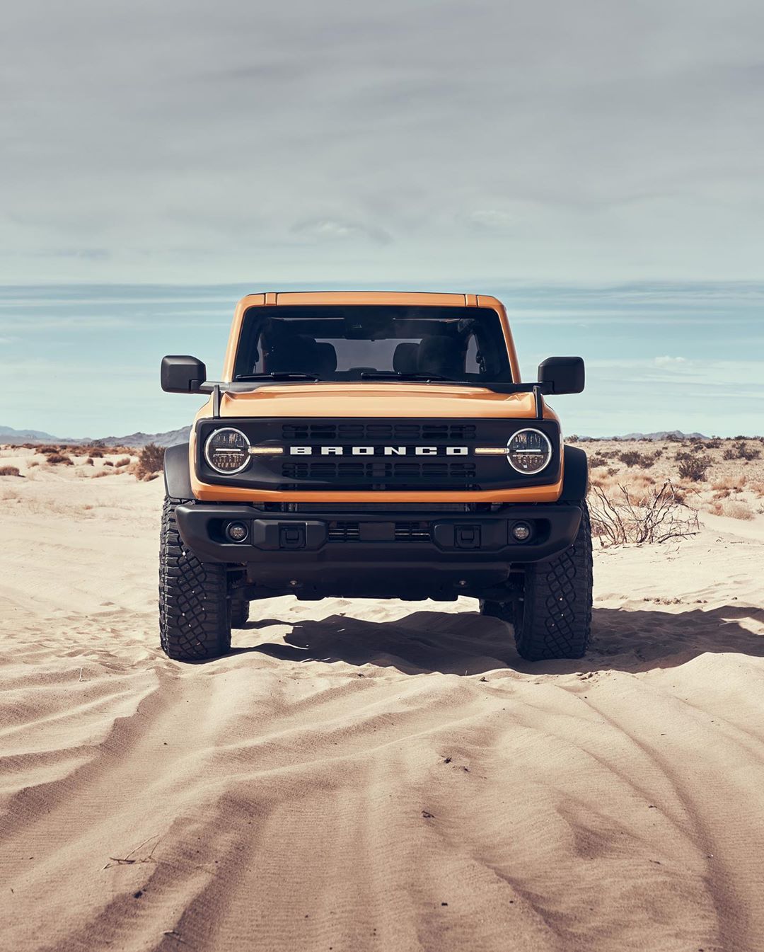 Front view of a yellow two-door Ford Bronco SUV parked on a sand dune against a partly cloudy sky in a desert landscape.