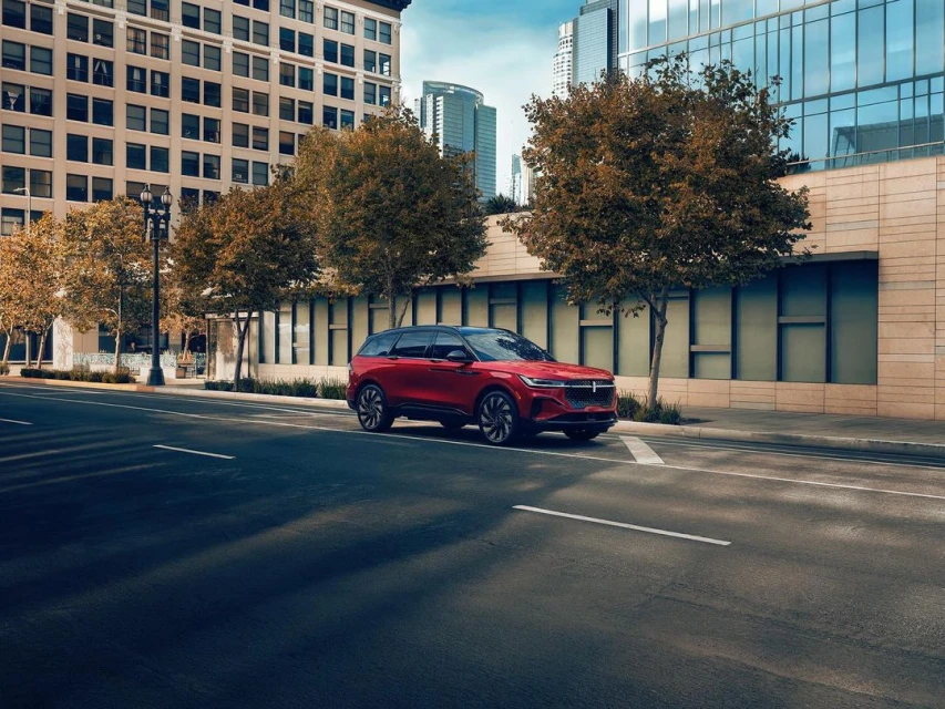 A red Lincoln Corsair SUV is parked on an urban street with tall buildings and trees in the background on a sunny day.