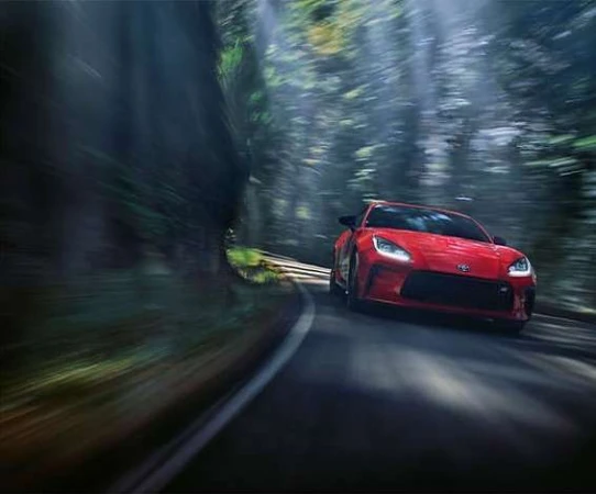 A red sports car drives along an asphalt road, blurred by motion, flanked by tall trees in a dense forest on a bright, sunny day.