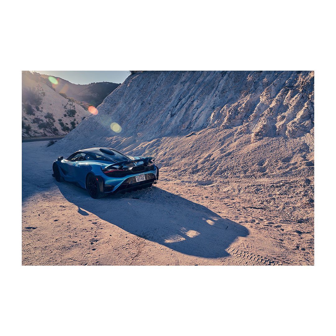 Blue sports car parked on a sandy surface with mountain in the background. The image captures automotive design and outdoor setting.