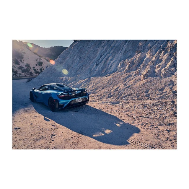 Blue sports car parked on a sandy surface with mountain in the background. The image captures automotive design and outdoor setting.