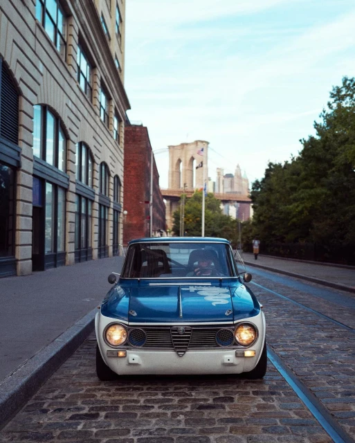 Front view of a blue and white Alfa Romeo parked on a cobblestone street with the Brooklyn Bridge in the background on a sunny day.