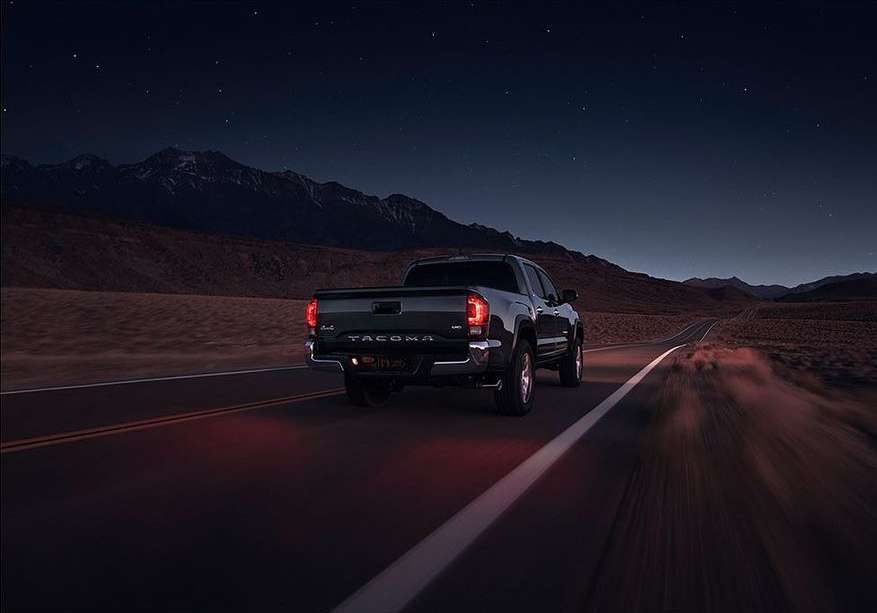 Black Toyota Tacoma driving on a dark desert road at night with mountain range and starry sky in the background. Automotive photography.