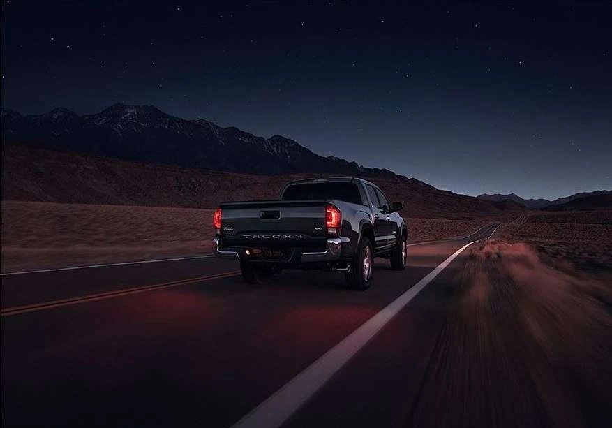 Black Toyota Tacoma driving on a dark desert road at night with mountain range and starry sky in the background. Automotive photography.