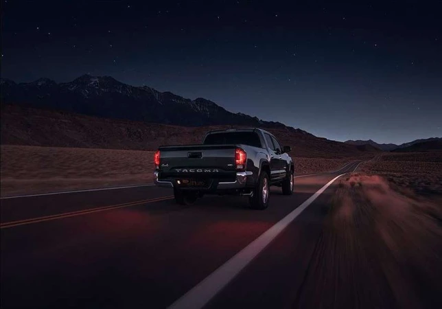 Black Toyota Tacoma driving on a dark desert road at night with mountain range and starry sky in the background. Automotive photography.