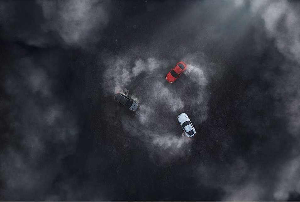Overhead shot of three cars drifting in a circle with tire smoke, creating a dynamic and dramatic scene on a dark surface.