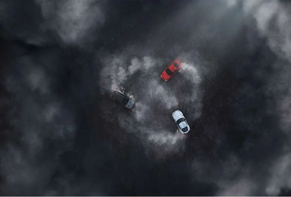 Overhead shot of three cars drifting in a circle with tire smoke, creating a dynamic and dramatic scene on a dark surface.