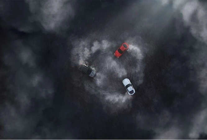 Overhead shot of three cars drifting in a circle with tire smoke, creating a dynamic and dramatic scene on a dark surface.