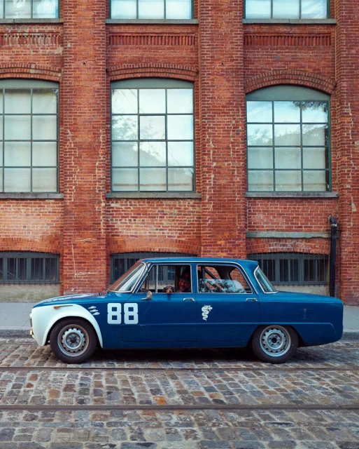 Vintage blue race car #88 parked on a cobblestone street in front of a brick building with window reflections.