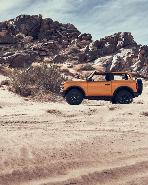 Yellow four-wheel drive vehicle parked in a sandy, desert landscape with rock formations and sparse vegetation under a cloudy sky.