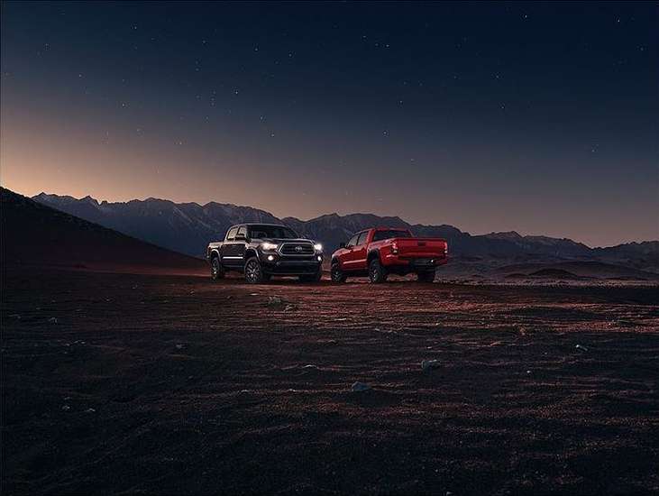 Two trucks, one black and one red, are parked in a desert landscape with mountains in the background against a twilight sky.