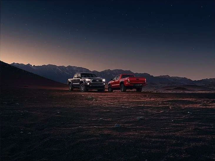 Two trucks, one black and one red, are parked in a desert landscape with mountains in the background against a twilight sky.