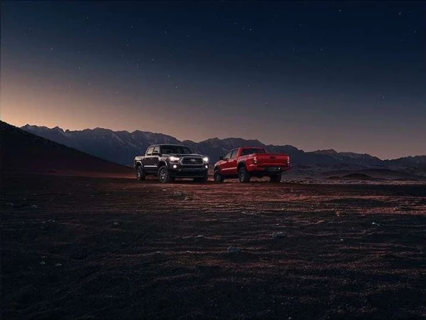 Two trucks, one black and one red, are parked in a desert landscape with mountains in the background against a twilight sky.