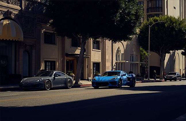 Two sports cars, one grey and one blue, are parked on a city street with trees and buildings on a sunny day, showcasing automotive design.