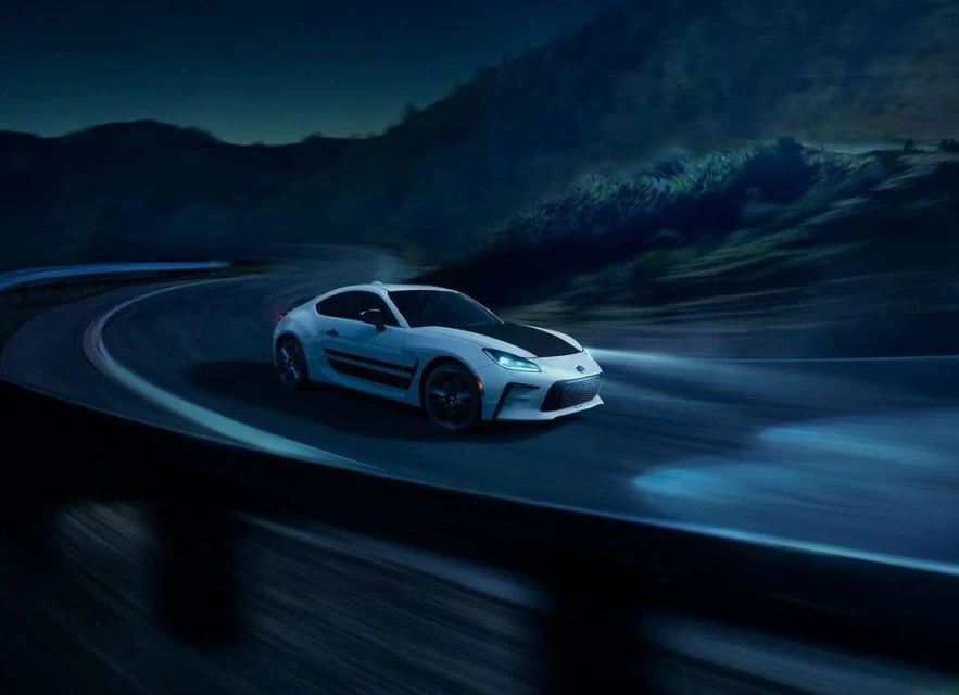 White Subaru BRZ driving on a winding road at night, with blurred motion, mountains in the background, showcasing automotive performance.
