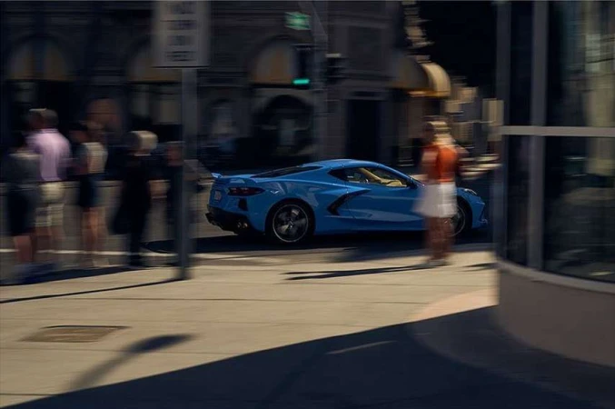 Powder-blue sports car driving on city street with pedestrians and building in the background. Blurred motion, urban setting, daylight.