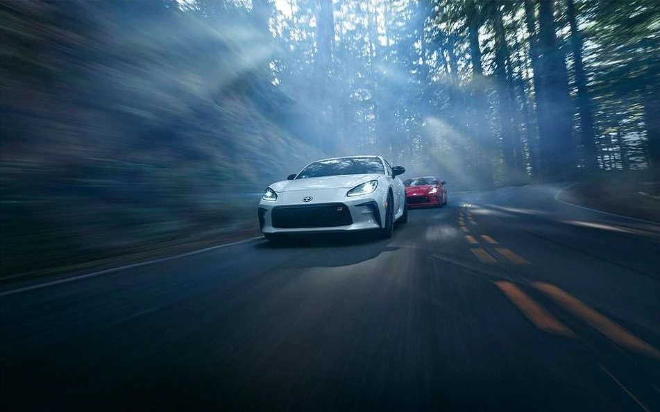 White sports car leads a red car driving fast through a forest with streaks of sunlight shining through the trees, creating motion blur.