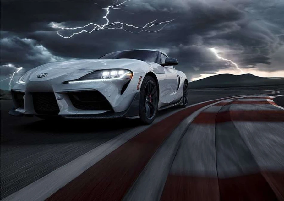 Sleek white Toyota Supra sports car speeding on a racetrack under a dramatic, stormy sky with visible lightning strikes.