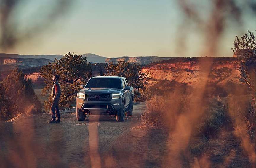 Truck parked on a dirt road in a desert landscape with a man standing nearby against a background of mesas and a sunset sky.