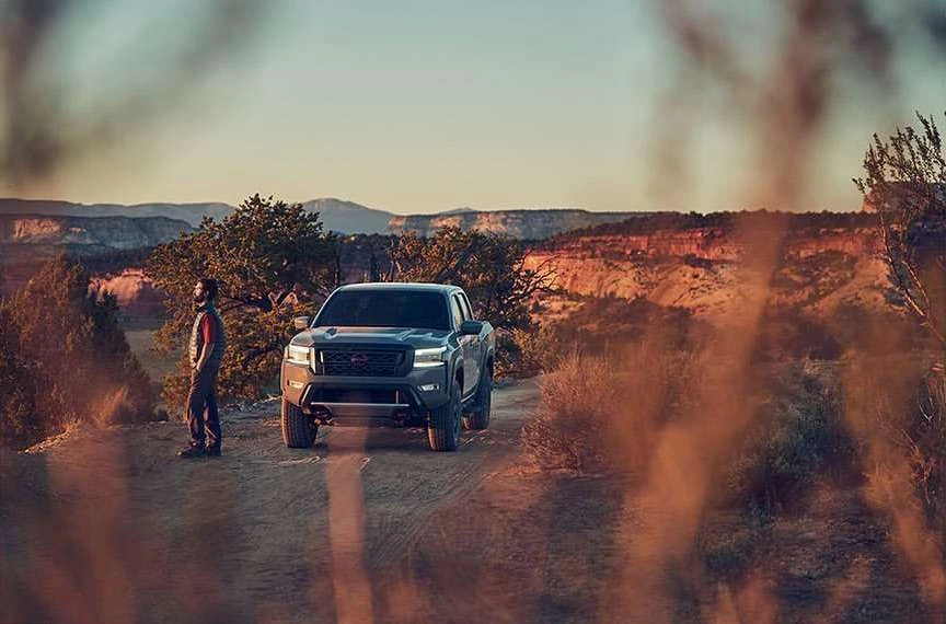 Truck parked on a dirt road in a desert landscape with a man standing nearby against a background of mesas and a sunset sky.