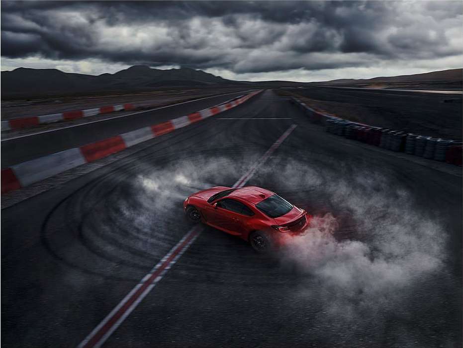 Red sports car drifting on a black asphalt race track with cloudy sky and mountain landscape in the background, creating a smoky effect.