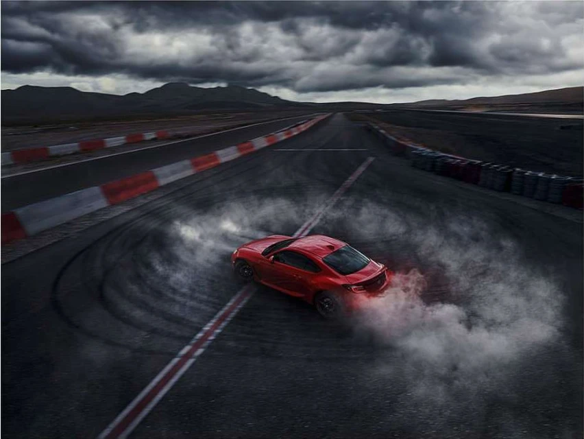 Red sports car drifting on a black asphalt race track with cloudy sky and mountain landscape in the background, creating a smoky effect.