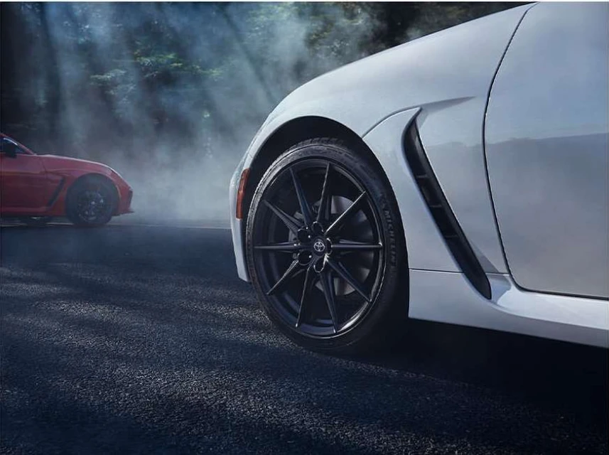White Toyota sports car wheel and fender in studio setting with red car in background and smoke effects, creating a dynamic car advertisement.